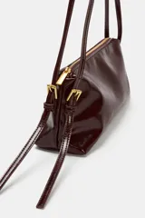 Bolso de hombro tipo baguette en color negro con acabado brillante. Presenta un diseño rectangular con cierre de cremallera superior y doble asa ajustable mediante hebillas metálicas.