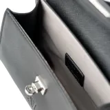 Cartera pequeña de mano tipo bandolera, confeccionada en símil cuero negro con textura saffiano. Presenta una solapa frontal con cierre de broche metálico plateado y el logo de la marca grabado en la parte inferior. Incluye asa superior fija y correa larga regulable y extraíble.