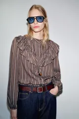 Camisa de manga larga con cuello subido, cierre frontal con botones y detalle de volantes en el pecho. Presenta un diseño a rayas verticales y puños elásticos.