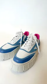 Zapatillas deportivas blancas con detalles en azul y rosa, cordones blancos y suela track.