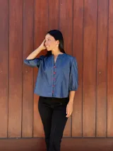 Camisa de jean azul con cuello mao, cierre frontal con botones rojos y mangas tres cuartos abullonadas con puños elásticos.