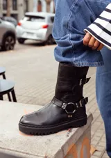 Bota biker de caña media confeccionada en cuero vacuno encerado. Presenta un diseño robusto con hebillas metálicas niqueladas en la caña y en la zona del tobillo, con una argolla decorativa. Cuenta con suela de goma negra con plataforma y cierre lateral interno.