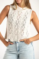 Top sin mangas de tejido de punto calado, color blanco, con cuello redondo acanalado y diseño de trenzas verticales. Presenta un ruedo con terminación festoneada.