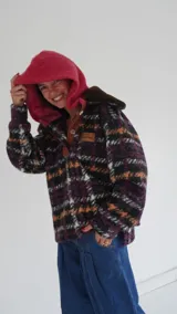 Buzo tipo canguro de lana con diseño oversize y estampado multicolor en tonos violeta, naranja y negro. Presenta una capucha amplia en color fucsia, cuello con botones y detalles en cuero.