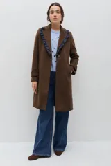 Tapado de lana color marrón con silueta entallada. Presenta solapas clásicas decoradas con un volado en contraste de color azul oscuro, cierre frontal con un botón y bolsillos laterales. Las mangas cuentan con detalles de botones en los puños.
