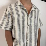 Camisa de manga corta con corte oversize, cuello tipo cubano y cierre frontal con botones. Presenta un diseño de rayas verticales en tonos crudo y gris.