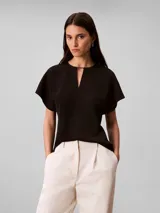 Blusa Calvin Klein de manga corta en color negro, confeccionada en tejido fluido de crêpe de chine. Cuenta con un diseño de cuello redondo con escote en V y una barra metálica decorativa en el centro del cuello.
