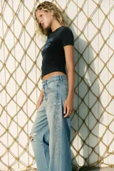 Jeans de tiro bajo y corte wide leg, confeccionados en denim de color celeste claro con lavado desgastado. Presentan detalles de roturas y aplicación de tachas metálicas en los laterales y bolsillos delanteros. Cierre frontal con cremallera y botón.