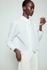 Camisa blanca de cuello solapa y manga larga, con detalle de lazada en el mismo tejido en la parte frontal y cierre con botones dorados.