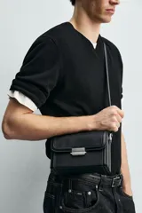 Bandolera rígida tipo caja, color negro, con solapa frontal y cierre metálico horizontal. Incluye asa de hombro ajustable.