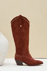 Bota texana de gamuza color marrón, con taco y puntera fina. Presenta bordados decorativos en el cuerpo de la caña y detalles de tachas metálicas en la parte superior y en la costura inferior.