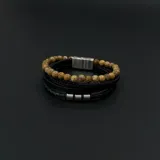 Pulsera multicapa para hombre que combina una hilera de cuentas de piedra natural en tonos beige con tres tiras de cuero negro, una de ellas trenzada y decorada con tres anillos metálicos. Incluye un cierre magnético de acero.
