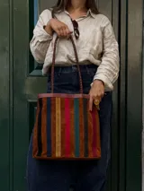 Cartera tipo tote de cuero con diseño de franjas verticales multicolores en tonos tierra, rojo y azul. Cuenta con correas trenzadas de cuero y cierre superior.