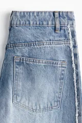 Jean de tiro alto, confeccionado en denim de algodón rígido, de corte ancho y pierna recta. Presenta un lavado celeste claro con efecto desgastado en el centro y franjas laterales en denim más oscuro con acabado desflecado.