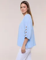 Blusa Erika de algodón con estampado de rayas en tonos azules, rosados y amarillos. Presenta cuello redondo con escote en V y botones frontales, mangas cortas decoradas con una fila de botones en los laterales y dobladillo asimétrico.