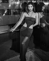 Vestido largo negro, ajustado al cuerpo, con escote strapless y tirantes finos cruzados en el cuello. Presenta recortes laterales pronunciados en la cintura y un nudo fruncido en la parte inferior frontal.