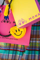 Llavero porta alcohol en gel con diseño de banana amarilla con cara sonriente. Incluye una correa de silicona negra, un dije metálico circular con frase grabada, un dije de silicona rosa y una letra M con glitter azul.