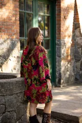 Vestido corto de lino con estampado floral en tonos fucsia, verde y negro. Presenta un diseño asimétrico, más corto en el frente y más largo en la parte posterior, con escote en V y mangas amplias.