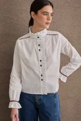 Camisa de popelina blanca con cuello alto tipo victoriano, volados frontales y detalles de costuras y ribetes en color marrón. Presenta mangas largas con puños elásticos terminados en volado y cierre frontal con botones oscuros.