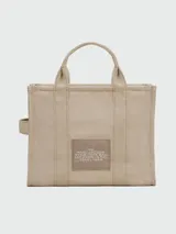Cartera tipo tote bag de Marc Jacobs, confeccionada en lona color beige con el logo estampado en blanco al frente. Cuenta con doble asa superior, correa de hombro ajustable y extraíble, y cierre superior con cremallera.