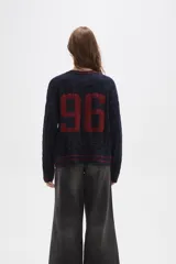 Sweater de tejido de punto con diseño de trenzas, cuello en V y detalles de rayas en contraste en cuello, puños y ruedo. Presenta un número bordado en el frente y tiene un corte holgado.