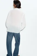 Blusa de tejido semitransparente con efecto arrugado, escote en pico y manga larga. Presenta un detalle decorativo frontal con estructura en forma de flor.