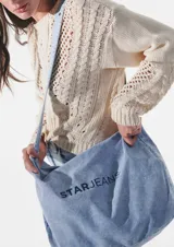 Bolso tipo tote de denim celeste con lavado ácido y estampado del logo "STARJEANS" en negro. Tiene correa larga regulable y cierre superior.