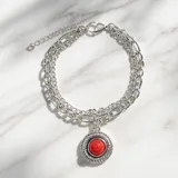 Pulsera de acero con cadena doble y dije redondo central que presenta una piedra roja tipo coral rodeada por un detalle trenzado.