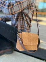 Cartera bandolera de cuero gamuzado color camel, con solapa frontal y herrajes metálicos en tono bronce. Incluye dos correas desmontables de cuero liso en tono marrón oscuro, una corta para llevar al hombro y una larga ajustable para usar cruzada.