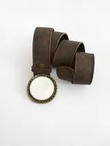 Cinto de cuero color chocolate oscuro con forro de cuero. Presenta una hebilla circular con borde metálico labrado y centro de guampa, con sistema de broche suizo intercambiable.