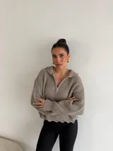Buzo de punto grueso color gris, con cuello polo, cierre frontal parcial y capucha. Presenta un diseño de tejido acanalado con terminaciones desflecadas en el ruedo.