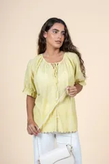 Blusa amarilla pastel de algodón con textura delicada, escote redondeado con lazo, mangas cortas abullonadas con volados y terminaciones sutiles.
