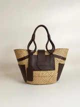 Bolso tipo tote confeccionado en rafia tejida y detalles de cuero marrón, con asas de mano y bolsillo frontal con solapa.