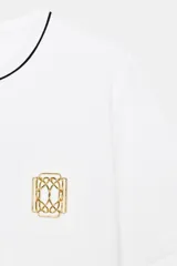 Camiseta negra de algodón, corte regular, con cuello redondo ribeteado en blanco y un bolsillo frontal con un broche metálico decorativo dorado.
