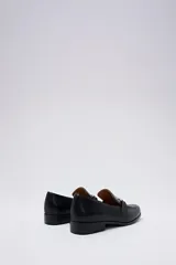 Mocasín de cuero negro con detalle de hebilla metálica plateada en el empeine y suela plana.