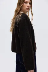 Chaqueta de sherpa suave en color marrón oscuro, con diseño de corte holgado y cuello redondo. Presenta ribetes de cinta finos en los bordes y cierre frontal mediante botones metálicos dorados.