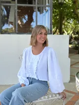 Blusa blanca de algodón con textura arrugada, cuello redondo, mangas largas abullonadas con puños elásticos y cierre frontal con botones y volantes.