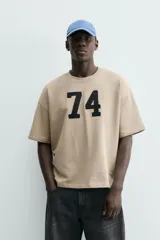 Sudadera de manga corta color beige, corte relajado (relaxed fit) con cuello redondo y número "74" bordado en negro en el frente.