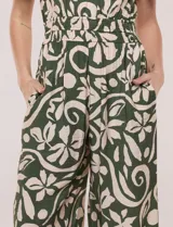 Enterito verde oscuro con estampado floral y abstracto en tono beige, de corte suelto y caída fluida. Presenta escote en V, mangas cortas, cintura elastizada con faja fruncida y pantalón crop de pierna ancha con bolsillos laterales.
