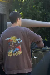Buzo marrón con estampa en la espalda de un sol, nubes, hongos y una persona sentada sobre flores.