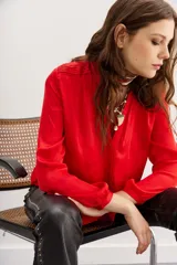 Camisa roja de manga larga con cuello redondo y escote frontal con botones ocultos. Presenta detalles de alforzas y frunces en los hombros.