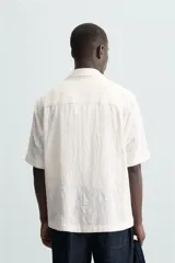 Camisa de corte relajado confeccionada en tejido de algodón con diseño jacquard floral. Presenta cuello bowling, manga corta, un bolsillo de parche en el pecho y aberturas laterales en el bajo. Cierre frontal con botones al tono.