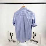 Camisa de vestir a rayas verticales azules y blancas, de corte clásico, con cuello italiano y mangas largas con puños abotonados. Presenta logo bordado en el pecho. Marca Duxon.