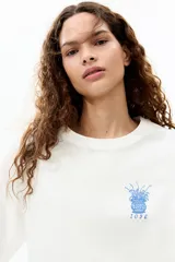 Remera oversized de algodón blanco, con cuello redondo acanalado, mangas cortas con hombros caídos y pequeño estampado azul en el pecho de un jarrón con flores y el número 1096.