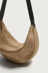 Bolso de hombro de gamuza marrón con diseño curvado, compartimento principal con cierre de cremallera y asa larga de cuero negro.