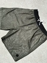 Short de baño verde oliva con estampado de letras y figuras geométricas en tono sobre tono. Cintura elástica negra con cordón blanco.