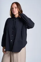 Camisa de color azul con cuello alto decorado con volados y lazo de ajuste. Presenta tablas decorativas en los hombros, cierre frontal con botones de nácar y mangas largas con puños elásticos que generan un efecto abullonado.