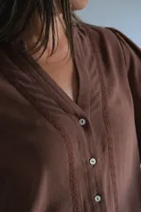 Blusa color chocolate con cuello en V, cierre frontal con botones y mangas largas abullonadas con puños elásticos. Presenta detalles de bordado lineal a lo largo de la abertura frontal.