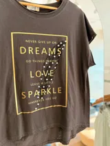 Remera de algodón color beige con corte relajado y diseño asimétrico, más larga en la parte posterior. Presenta un estampado frontal rectangular con letras doradas, una pluma ilustrada y detalles de pedrería decorativa.