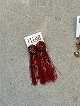 Caravanas colgantes artesanales FLUSS, con base circular de resina color bordó incrustada con detalles dorados y rojos, de la cual cuelgan largos flecos de hilo rojo, cadenas doradas y pequeños dijes de piedra y cuentas.
